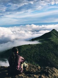 Rear view of woman looking at view while sitting on mountain against cloudy sky