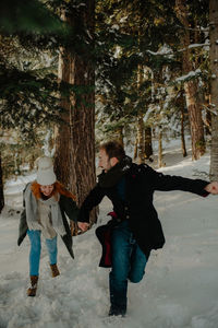 Full length of couple holding hand running on snow covered land in forest