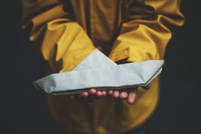 Midsection of girl holding paper boat while wearing yellow raincoat