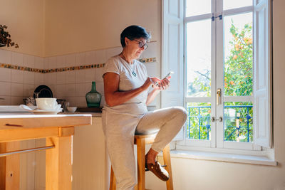 Mature female in casual clothes and glasses sitting on stool near window and browsing mobile phone in cozy home kitchen with wooden furniture in daylight