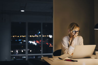 Dedicated mature businesswoman using laptop while sitting at illuminated desk in coworking space