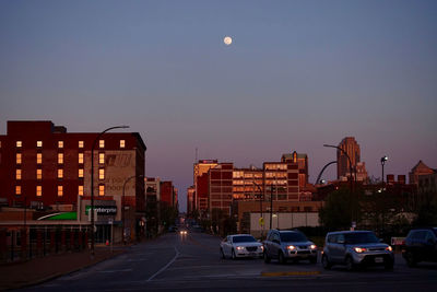 Cars on road by buildings against sky at dusk