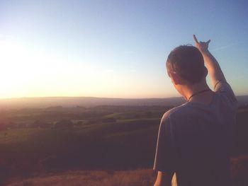 Rear view of man with horn sign against landscape during sunset