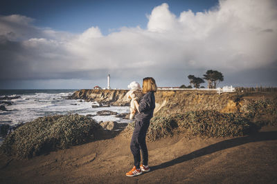 A woman is holding a baby near a lighthouse on the pacific coast