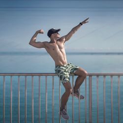 Full length of shirtless man sitting on railing against sky