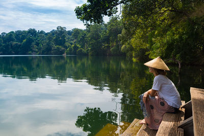 Rear view of man sitting by lake against trees