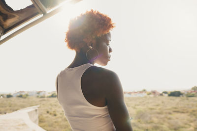 Back view of confident thoughtful african american female with dyed hair in trendy outfit standing near weathered structure on sunny summer day on street