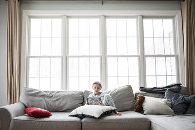 Wide view of girl home sick from school on couch with tablet and teddy