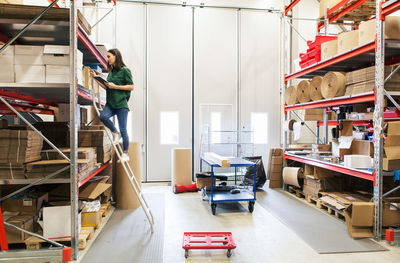 Woman analyzing cardboard box while standing on ladder in warehouse