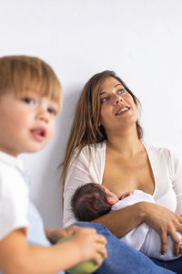 Contemplative mother breastfeeding newborn daughter by son at home