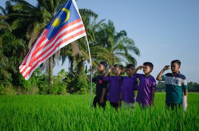 Boys saluting while standing by malaysian flag on field against clear sky