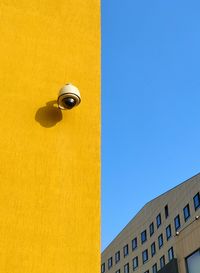 Low angle view of security camera on yellow building against blue sky