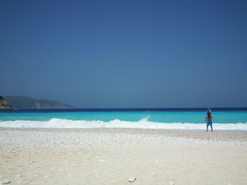 Man carrying child while walking on shore at beach against clear blue sky