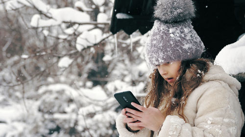 Dressed in warm winter clothes fully covered with snowflakes is sitting on bench and using smartphon