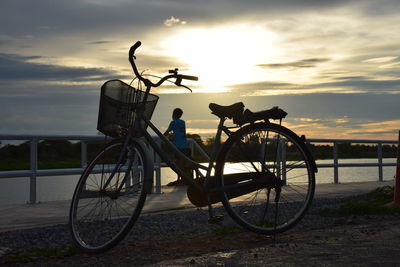 Bicycle parked by railing against sky during sunset