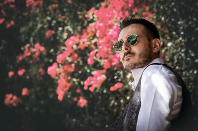 Side view of young man wearing sunglasses standing against red flowering plants