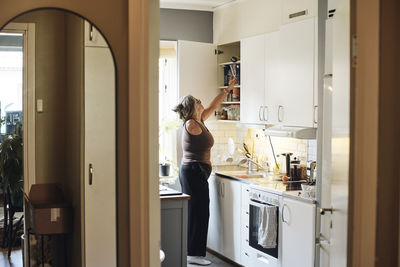 Side view of mature woman with disability searching in cabinet while standing in kitchen at home