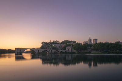 Bridge over river by buildings against sky during sunset