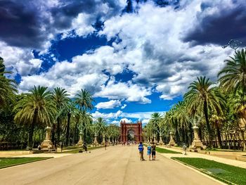 People walking on footpath by palm trees and buildings against sky