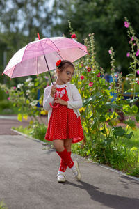 A beautiful little girl in a red dress and white blouse under a pink umbrella stands on a summer day