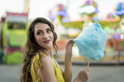 Side view of happy female eating sweet blue cotton candy while having fun and enjoying weekend at fairground in summer
