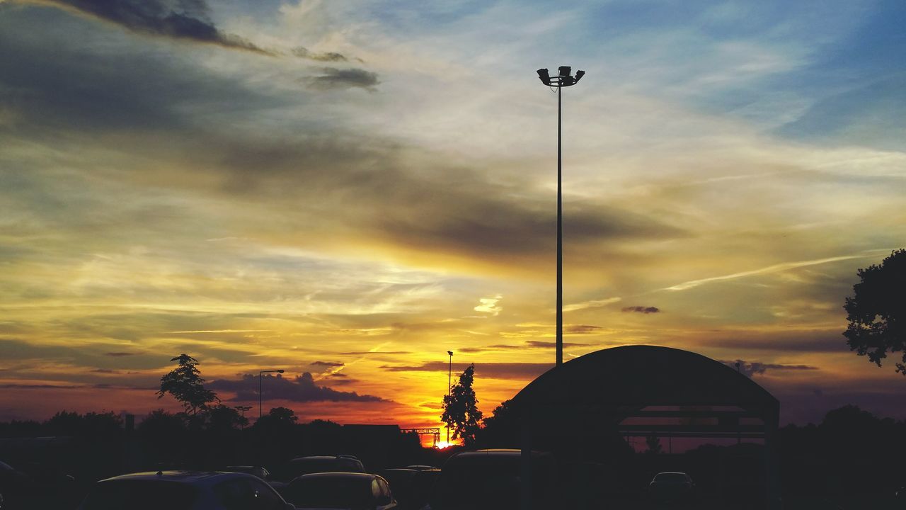 sunset, car, land vehicle, transportation, sky, mode of transport, street light, cloud - sky, silhouette, orange color, street, cloudy, road, dramatic sky, cloud, building exterior, lighting equipment, parking, tree, beauty in nature