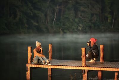 Woman photographing female friend sitting on pier in lake