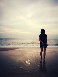 Rear view of young woman standing on sea shore against cloudy sky during sunset