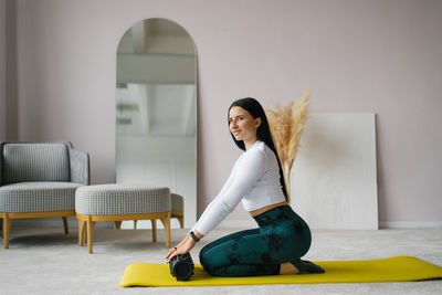 Young woman holds a roller for myofascial release in her hands, sitting at home on a gym mat