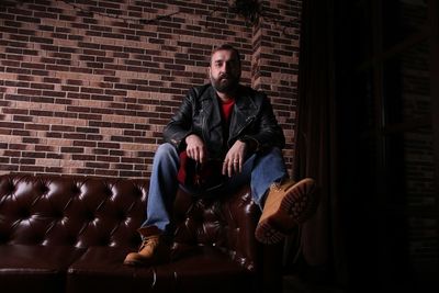 Full length of fashionable man sitting on sofa against brick wall