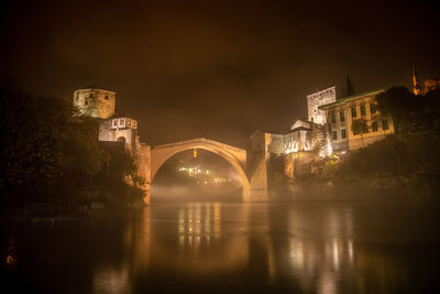 Illuminated bridge over river against buildings at night