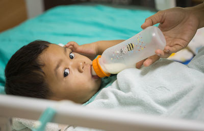 Cropped hands of parent feeding milk to son lying on bed in hospital