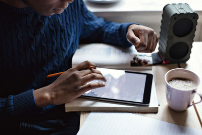 Midsection of teenage boy using digital tablet by coffee cup on table while doing homework