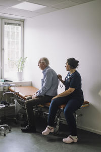 Female nurse with stethoscope examining senior male patient sitting on bed in clinic