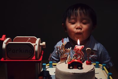 Cute boy with eye closed blowing birthday candle on cake in darkroom