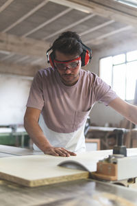 Young male carpenter cutting wood using table saw while working in workshop