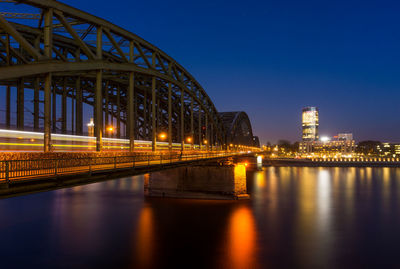 Illuminated bridge over river by buildings against sky at night