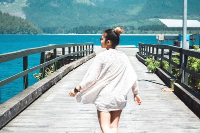 Rear view of woman walking on boardwalk by sea against mountains on sunny day