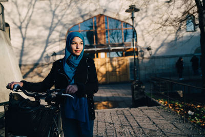 Confident young woman walking with bicycle on footpath against building in city