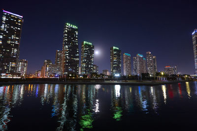 Illuminated buildings by river against sky at night