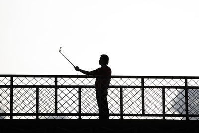 Silhouette man photographing on footbridge against clear sky