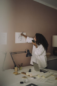 Busy female architect talking through speaker phone while examining blueprint at office