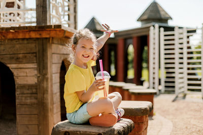 The girl is sitting on the playground and drinking juice while playing