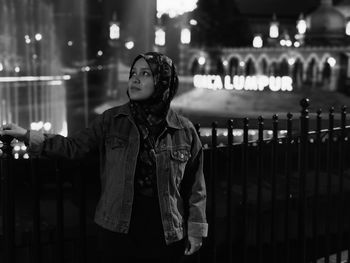 Woman wearing hijab looking away while standing by fence in city at night