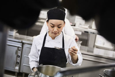 Young female chef tasting food from cooking pan at commercial kitchen