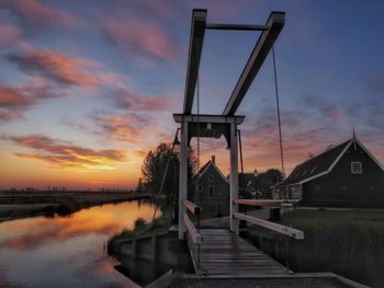 Footbridge by river against sky during sunset