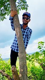 Portrait of young man standing by tree against sky