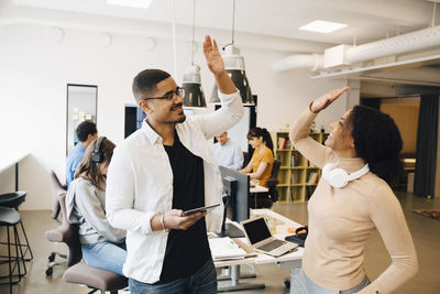 Smiling programmers doing high-five while standing with colleagues working in background