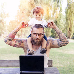 Portrait of bearded man using laptop while son sitting on shoulder in park