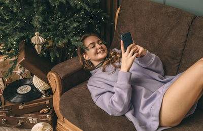 Young beautiful girl lies near decorated christmas tree on sofa, smiling and using phone,  fir xmas